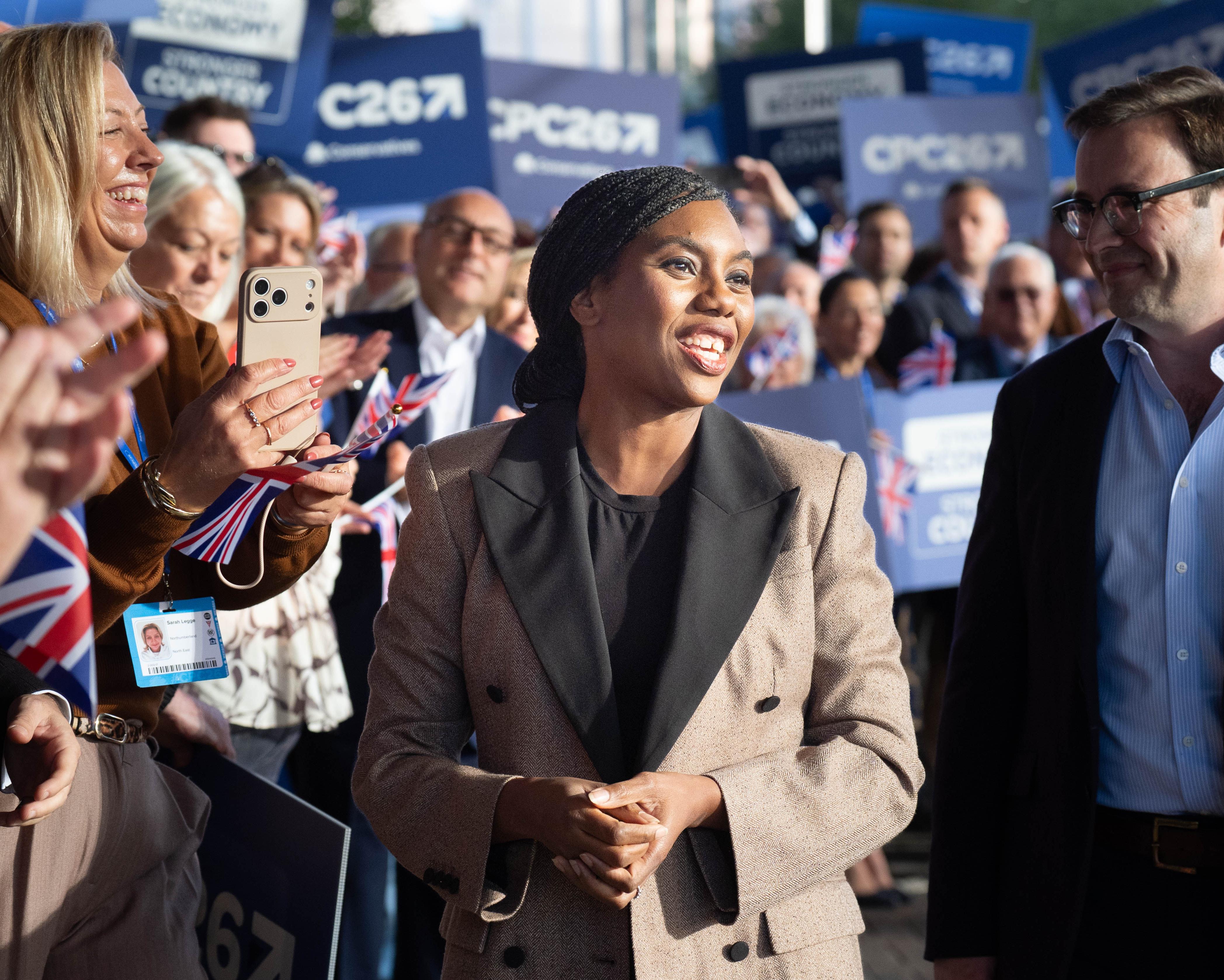 A smiling woman in a beige blazer walks past supporters holding Conservative signs and Union Jack flags.