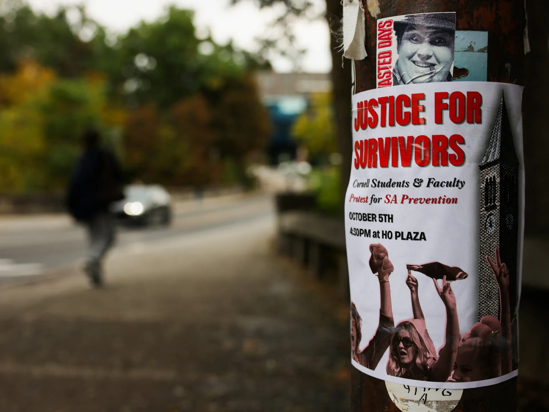 A campus pole bears a “Justice for Survivors” protest flyer, with a blurred pedestrian and street behind it.