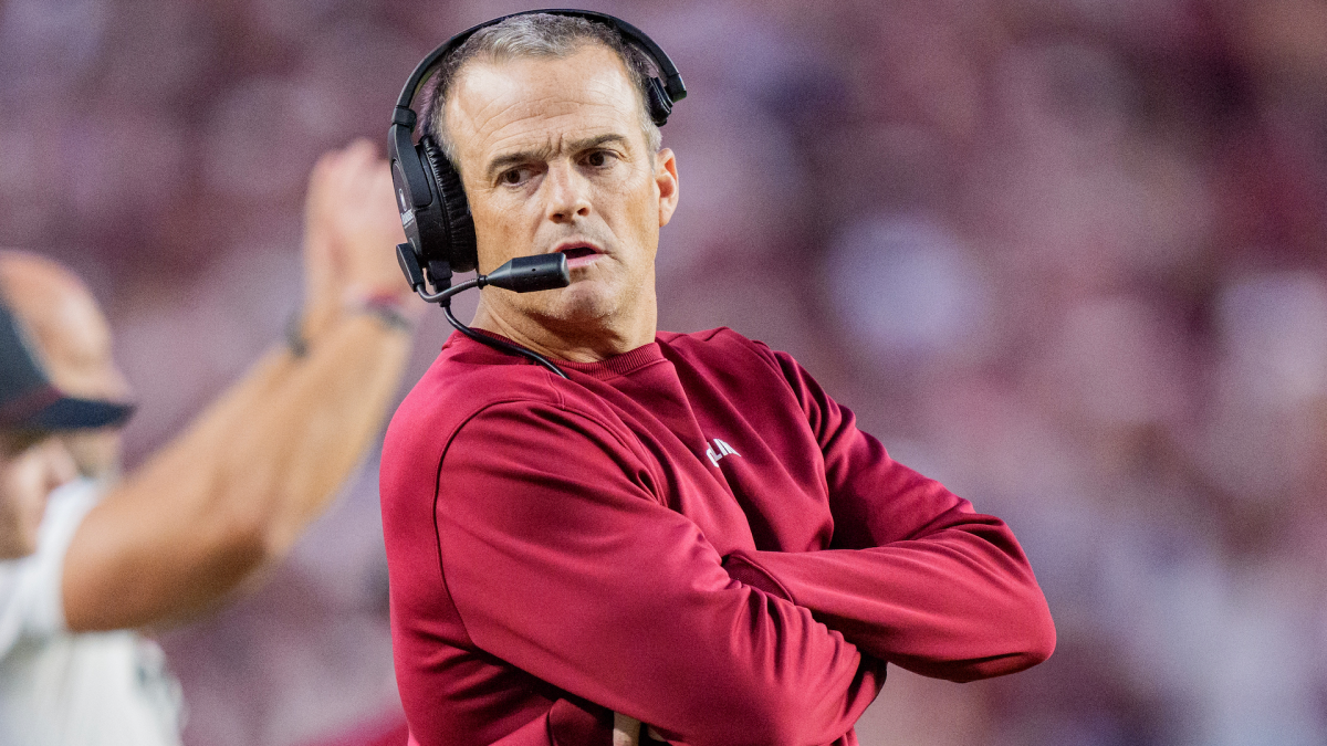 A man in a garnet sweatshirt and headset stands with arms crossed against blurred stadium seating.