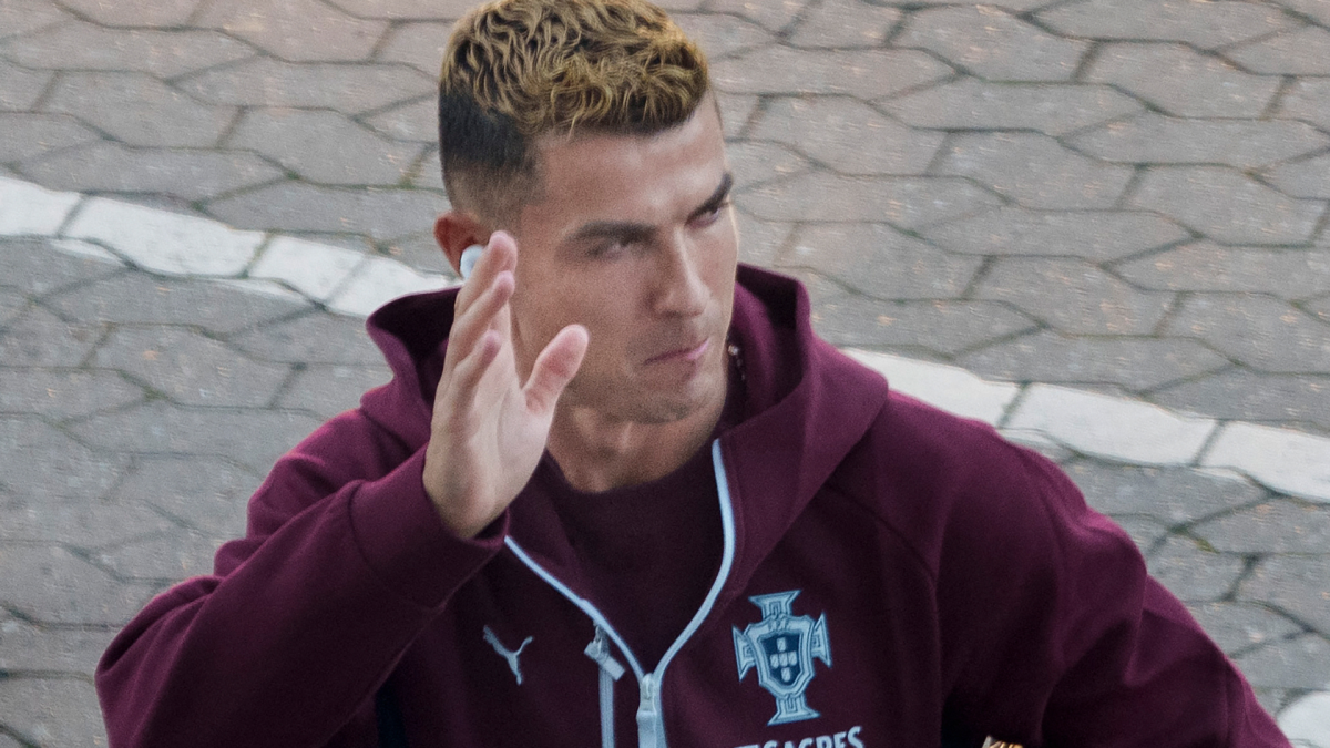 Man in a burgundy Portugal jacket raises one hand while walking across stone paving.