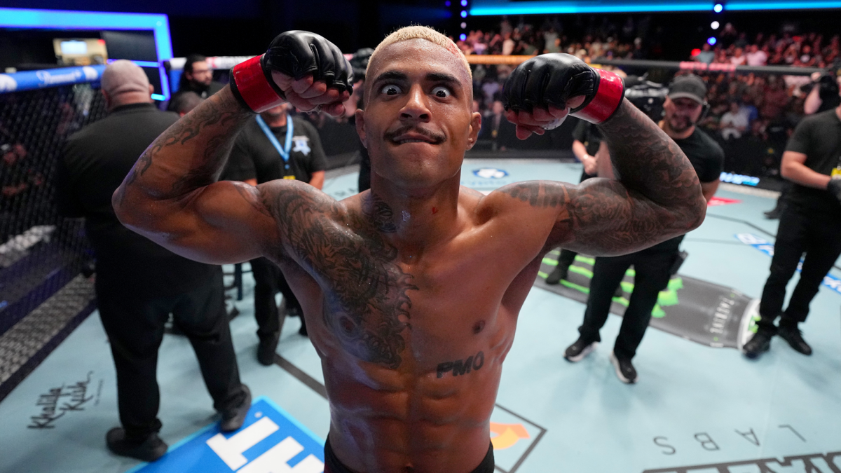 Tattooed fighter flexes both arms inside an MMA cage, wearing black gloves with red wrist wraps.