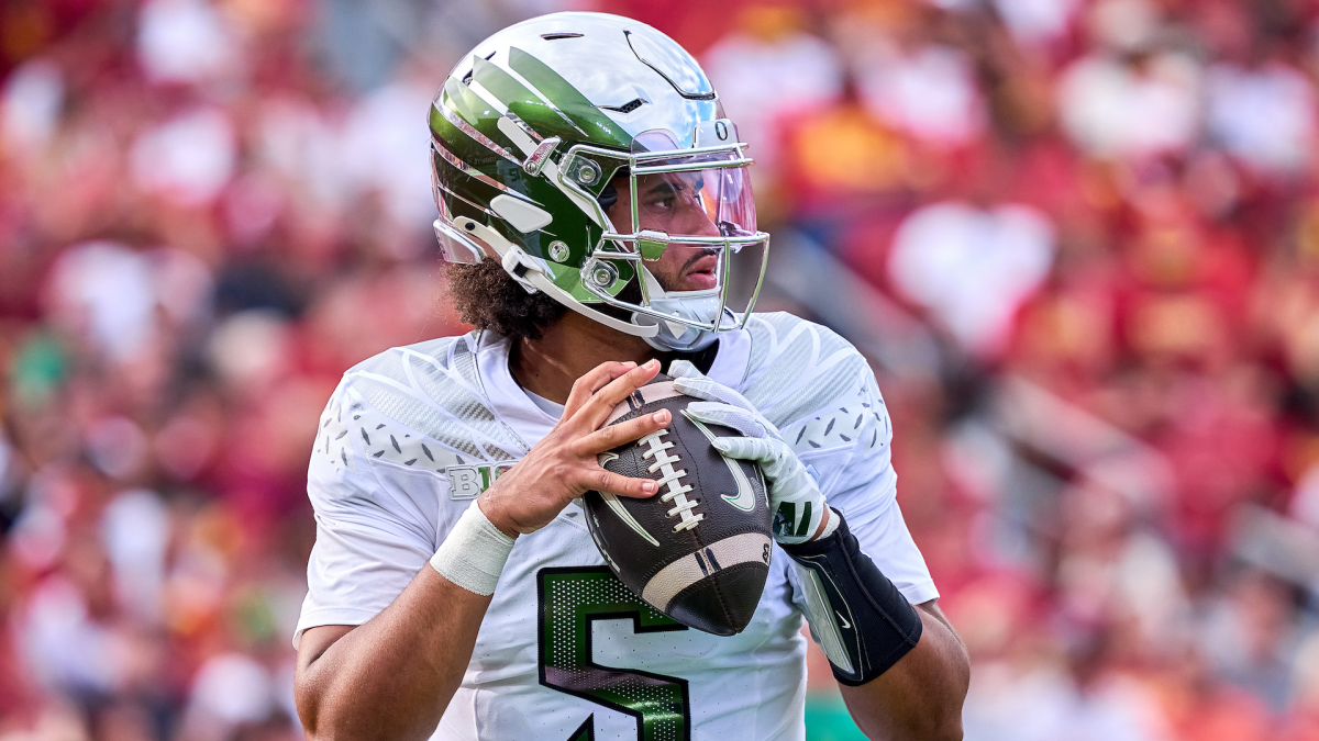 Football player in a white uniform holds the ball, looking downfield against a blurred crowd.