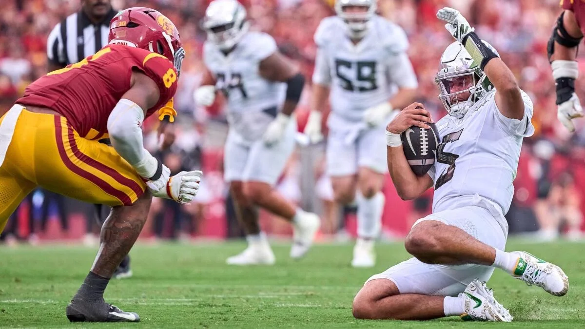Oregon quarterback in white raises his arm while holding the football as a USC defender approaches.