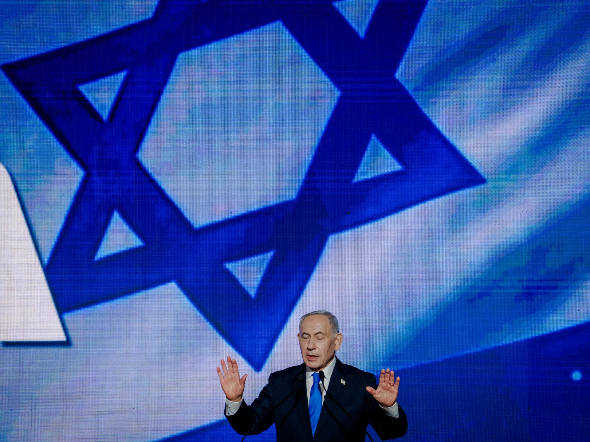 A suited speaker gestures at a podium beneath a large blue Star of David graphic.