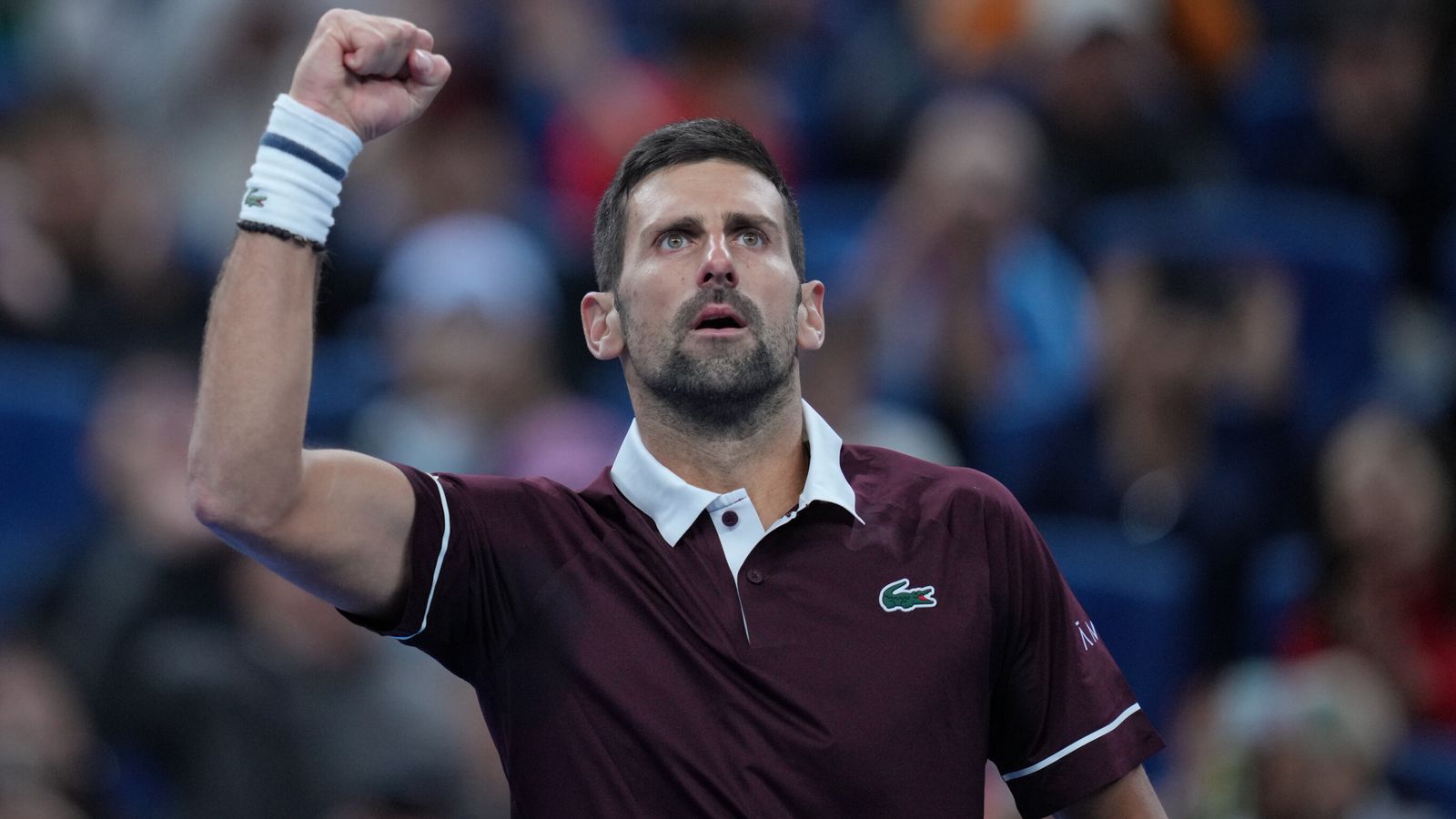Tennis player in a burgundy shirt raises a clenched fist before a blurred crowd.