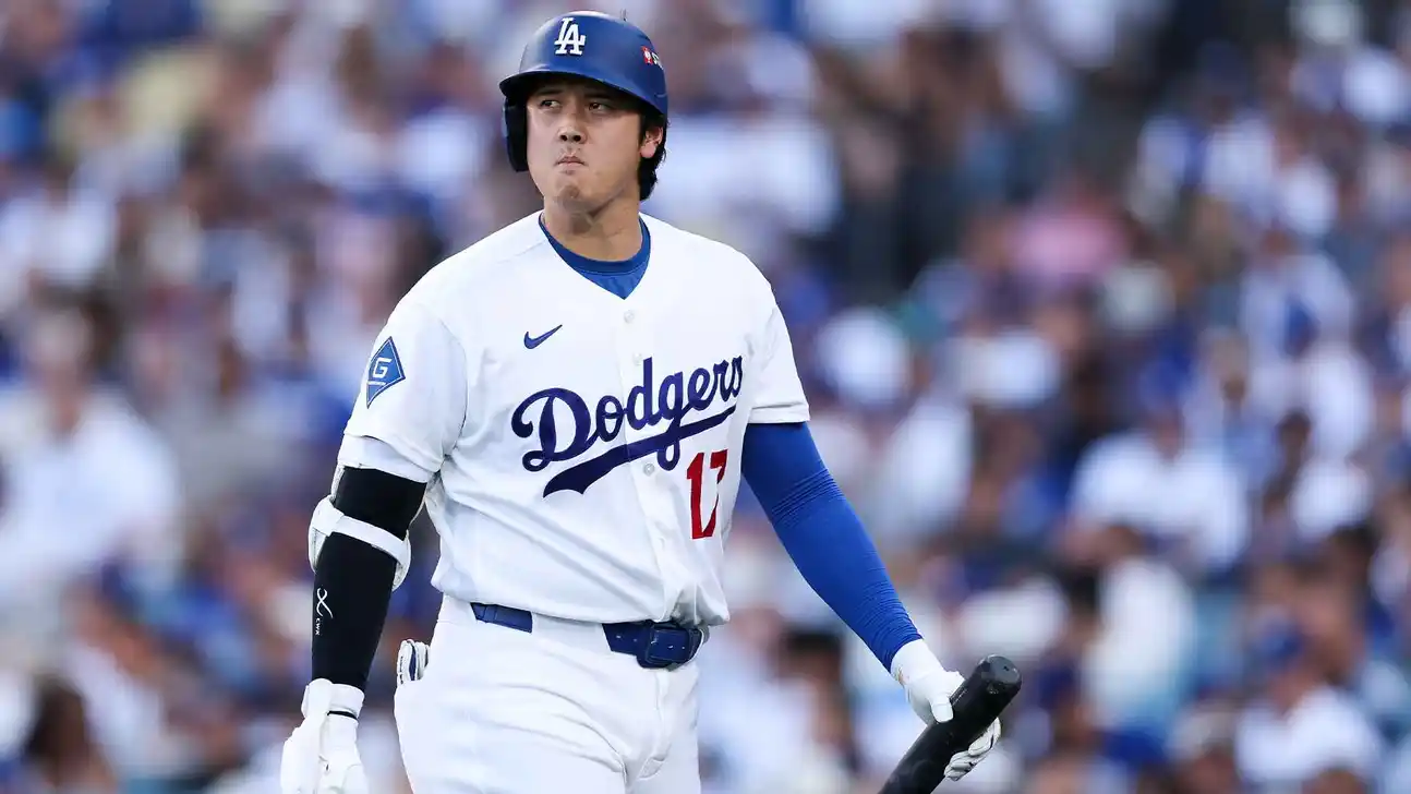 Dodgers player in a white uniform and blue helmet walks onto the field holding a bat.