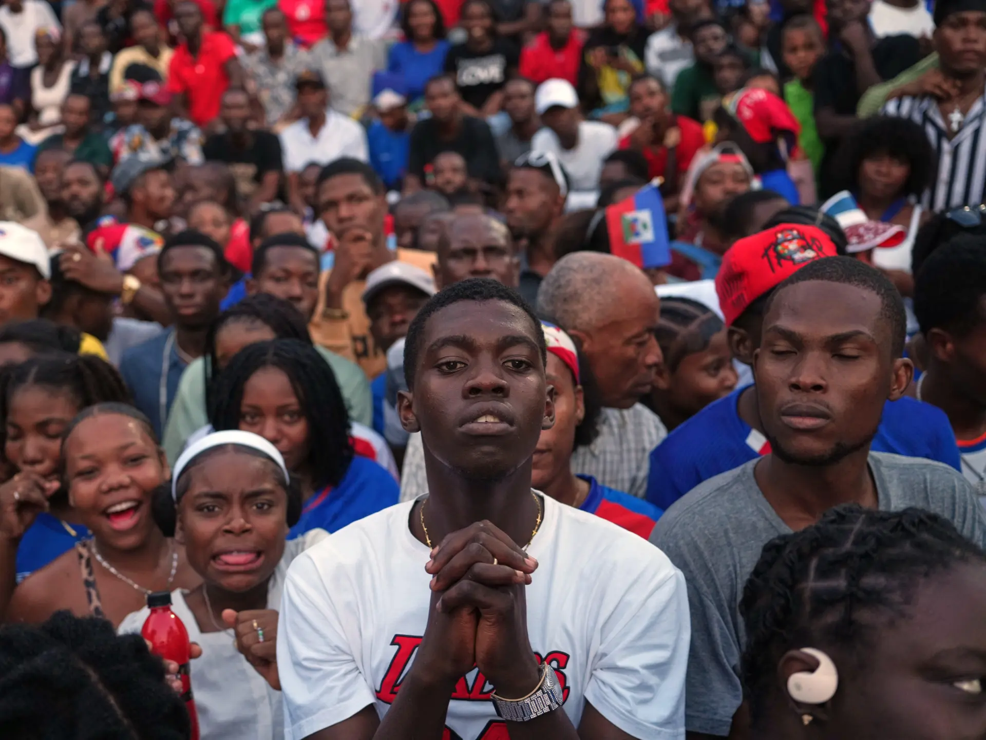 A young man clasps his hands amid a crowded group of football fans, some holding Haitian flags.
