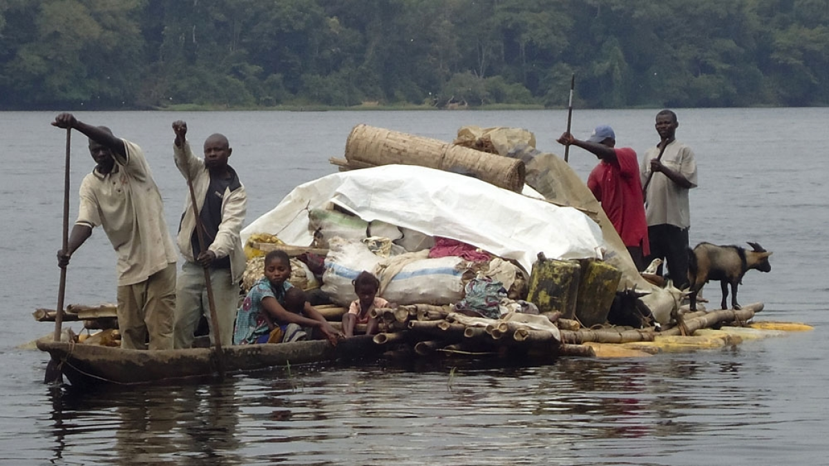People, children, and goats ride a heavily loaded raft across a broad river.