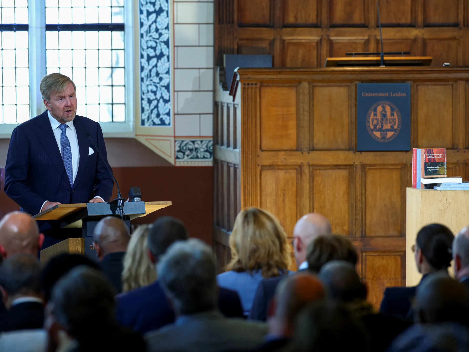 A suited speaker addresses an audience from a lectern in a wood-paneled hall.