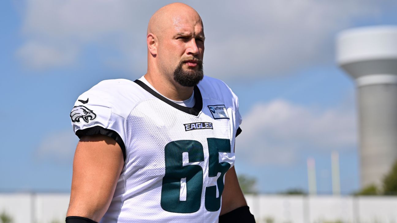 Bald, bearded football player in a white Eagles jersey stands outdoors beneath a blue sky.