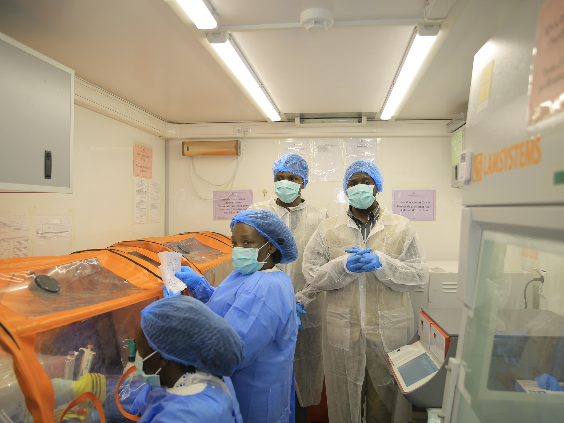 Healthcare workers in protective clothing stand beside orange isolation equipment in a clinical laboratory.