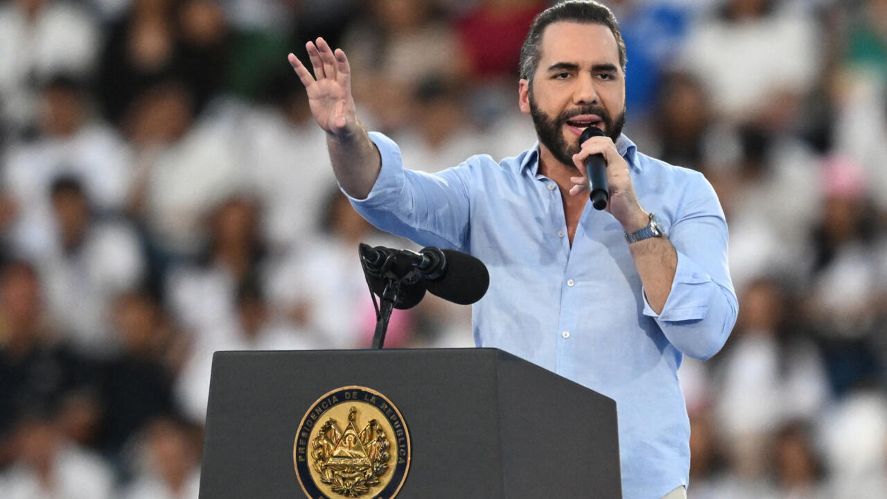 A man in a light blue shirt speaks into a microphone behind a dark podium, raising one hand.