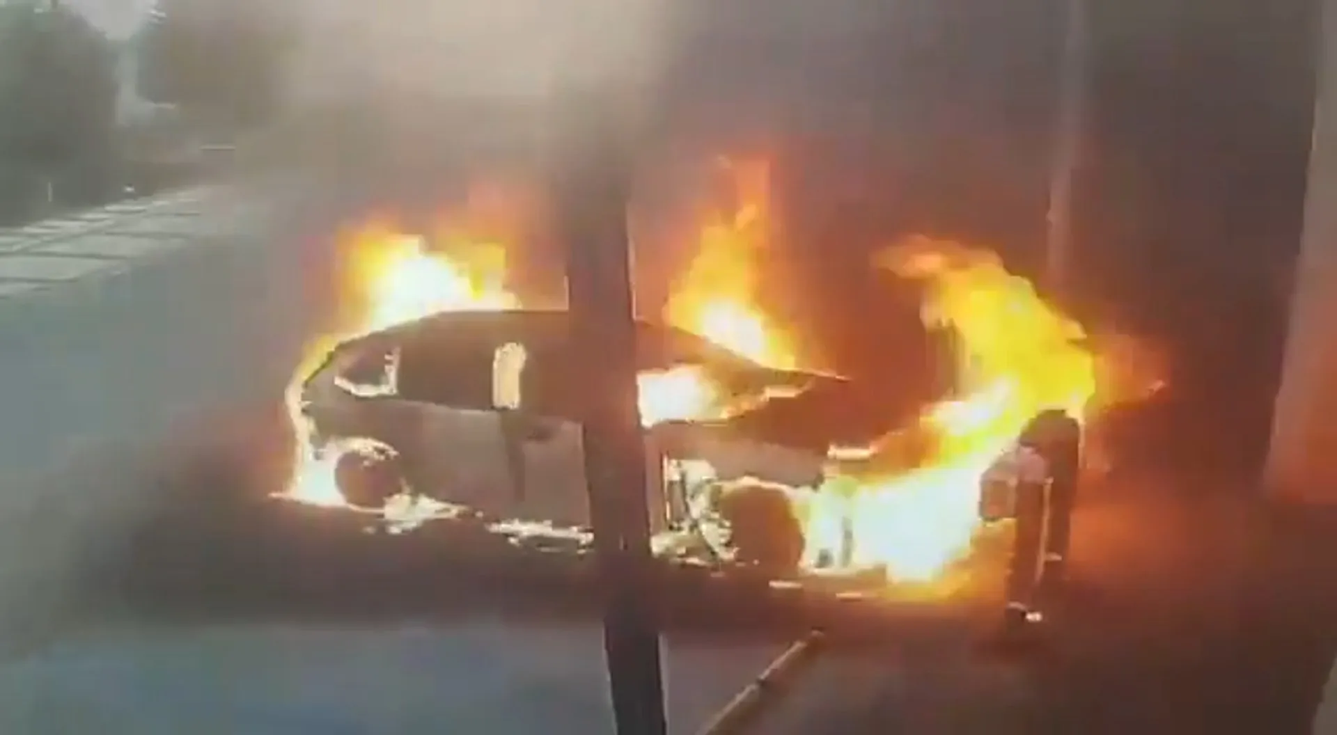 A car burns beside a charging station as flames rise around its front and side.
