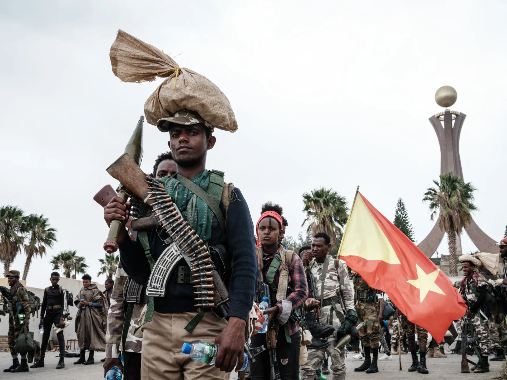 Armed men gather in a public square, one carrying a rifle and another holding a red and yellow flag.