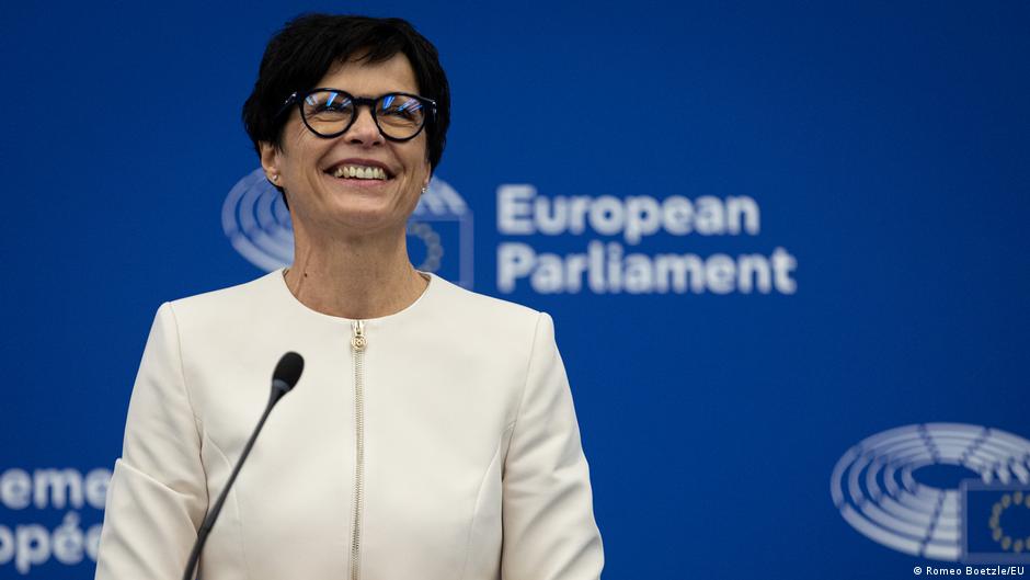 A smiling woman in glasses and a white jacket stands at a microphone before a European Parliament backdrop.