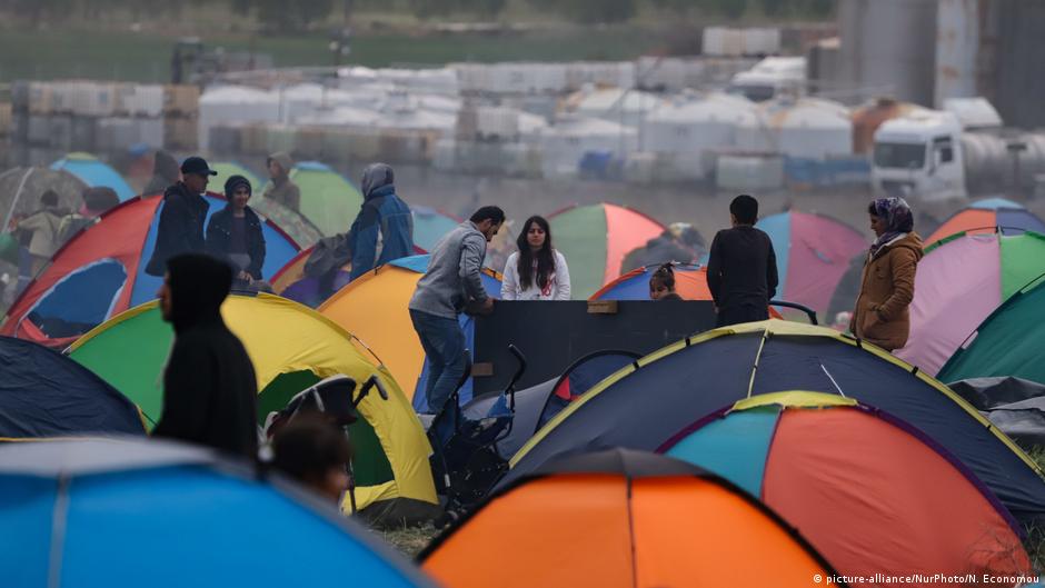 People stand among colorful tents at a crowded outdoor encampment, with vehicles and barriers in the background.