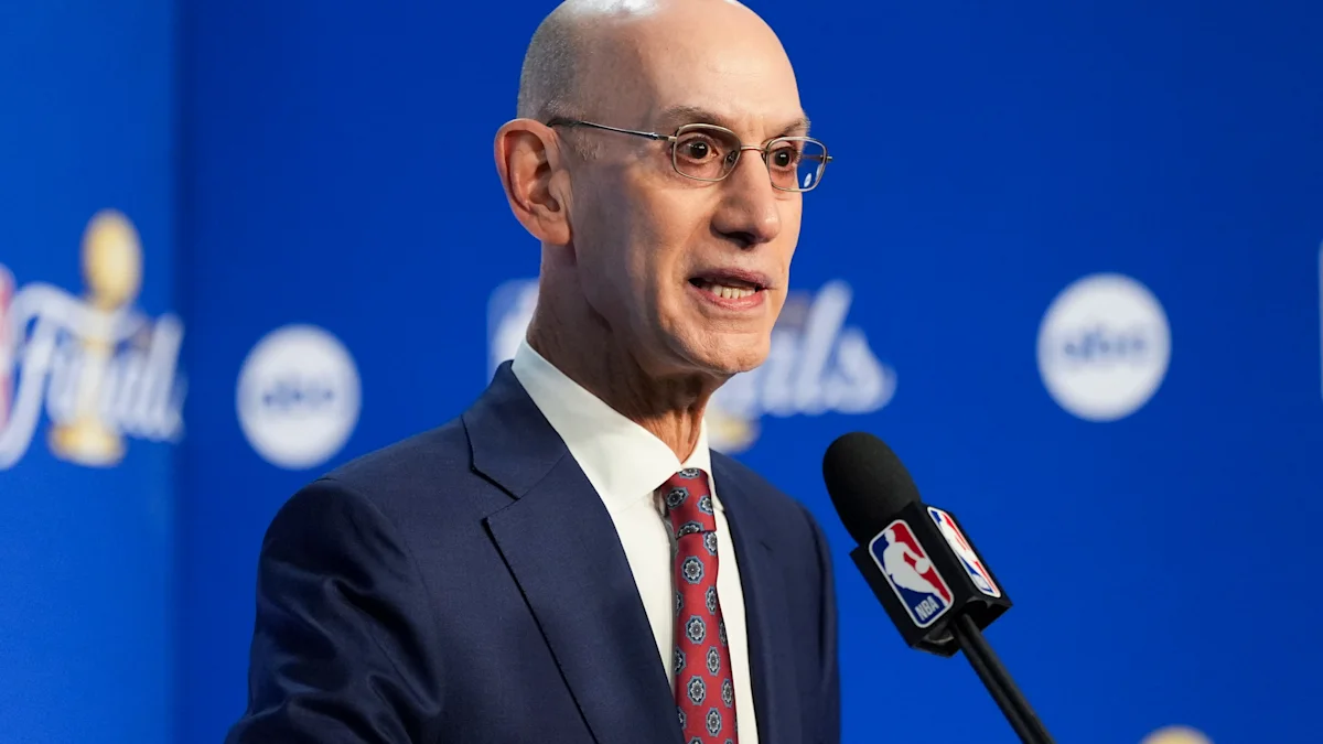 A bald man in glasses and a navy suit speaks into an NBA-branded microphone against a blue backdrop.