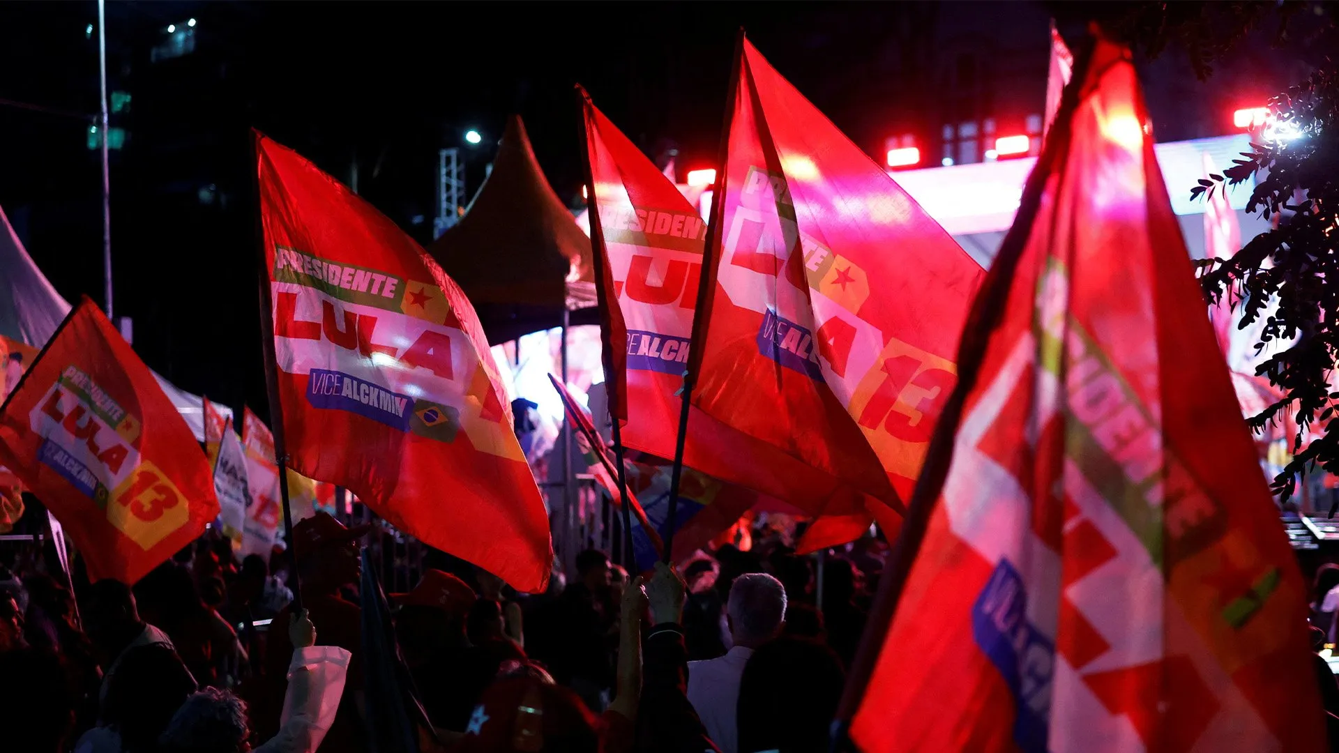 Red campaign flags wave above a crowd at a brightly lit nighttime political rally.