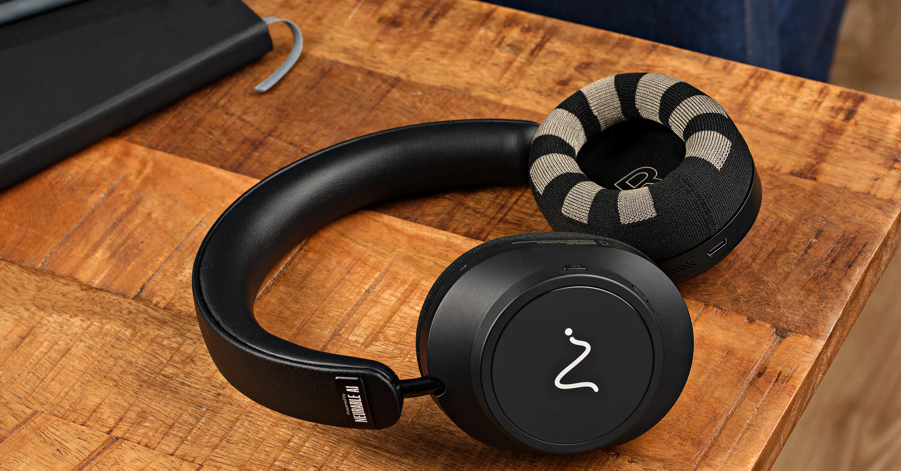 Black over-ear headphones with a striped fabric ear cushion rest on a wooden table.