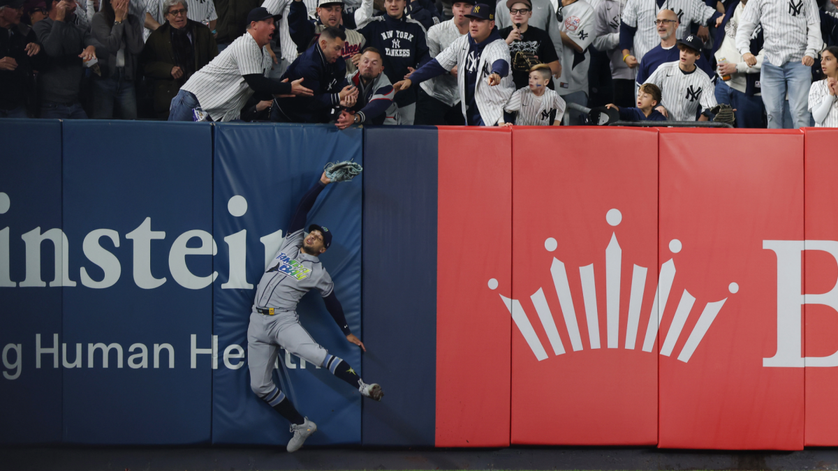 A Rays outfielder leaps at the wall as Yankees fans reach over from the stands.