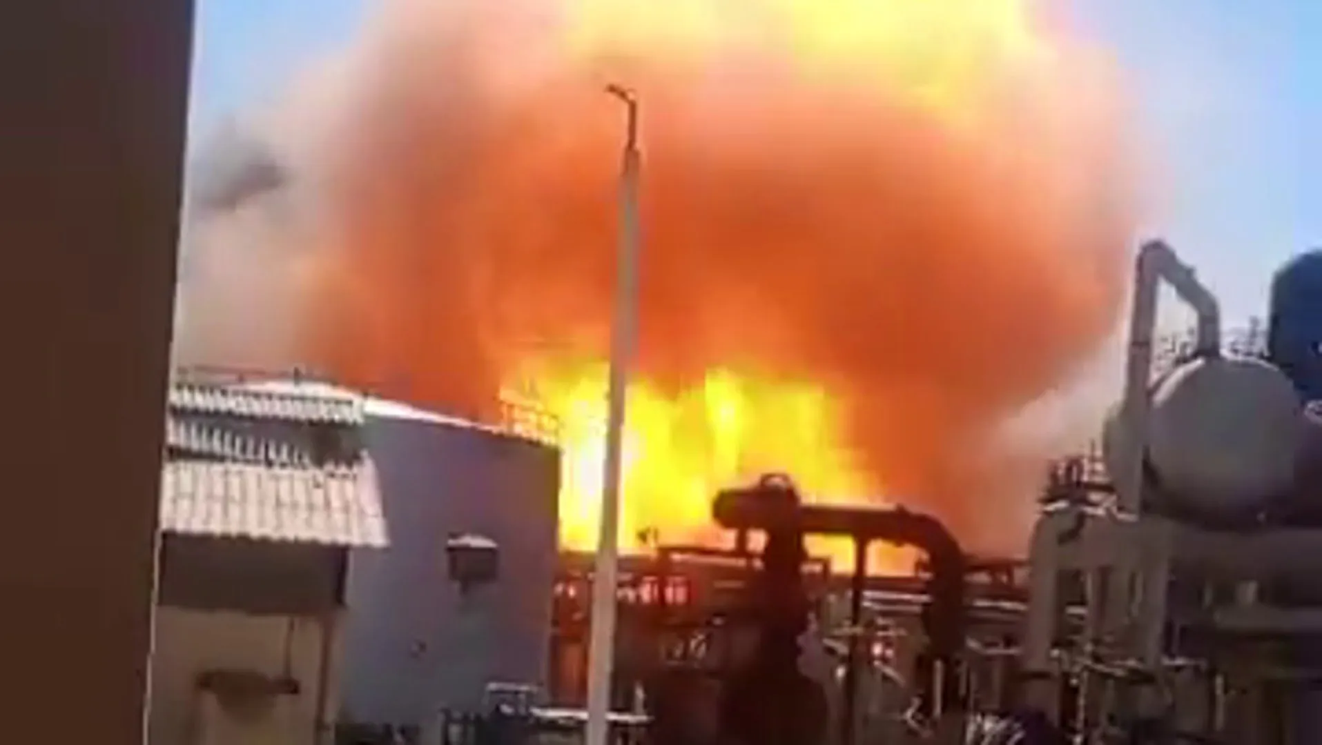 Bright flames and thick orange smoke rise behind industrial equipment and nearby buildings.