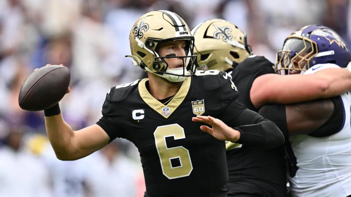 Saints quarterback in a black number 6 jersey prepares to throw as teammates block a Ravens defender.