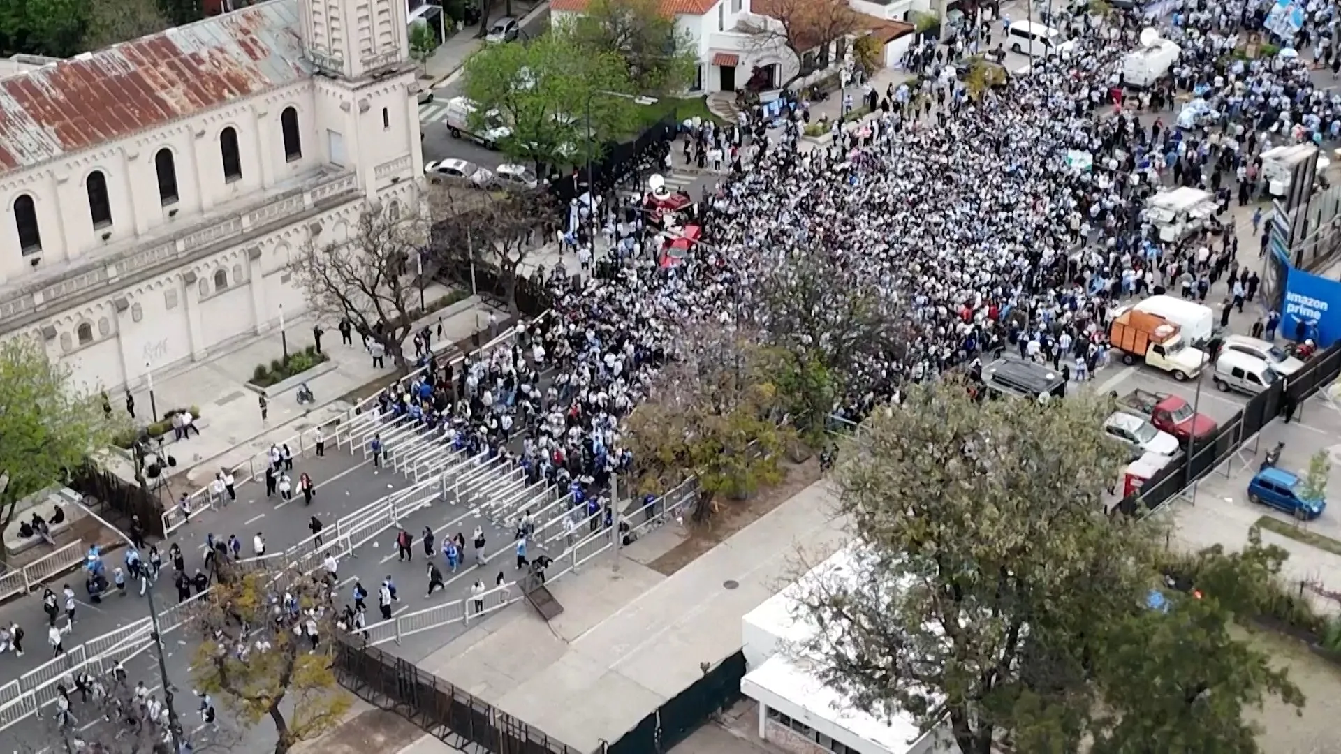 Aerial view of a dense crowd filling a street beside a large building and metal barriers.