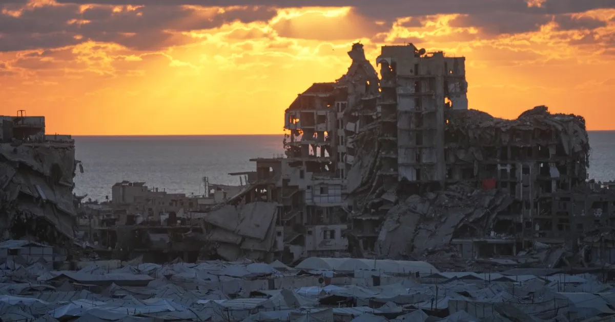A ruined coastal city at sunset, with damaged buildings and rows of tents in the foreground.