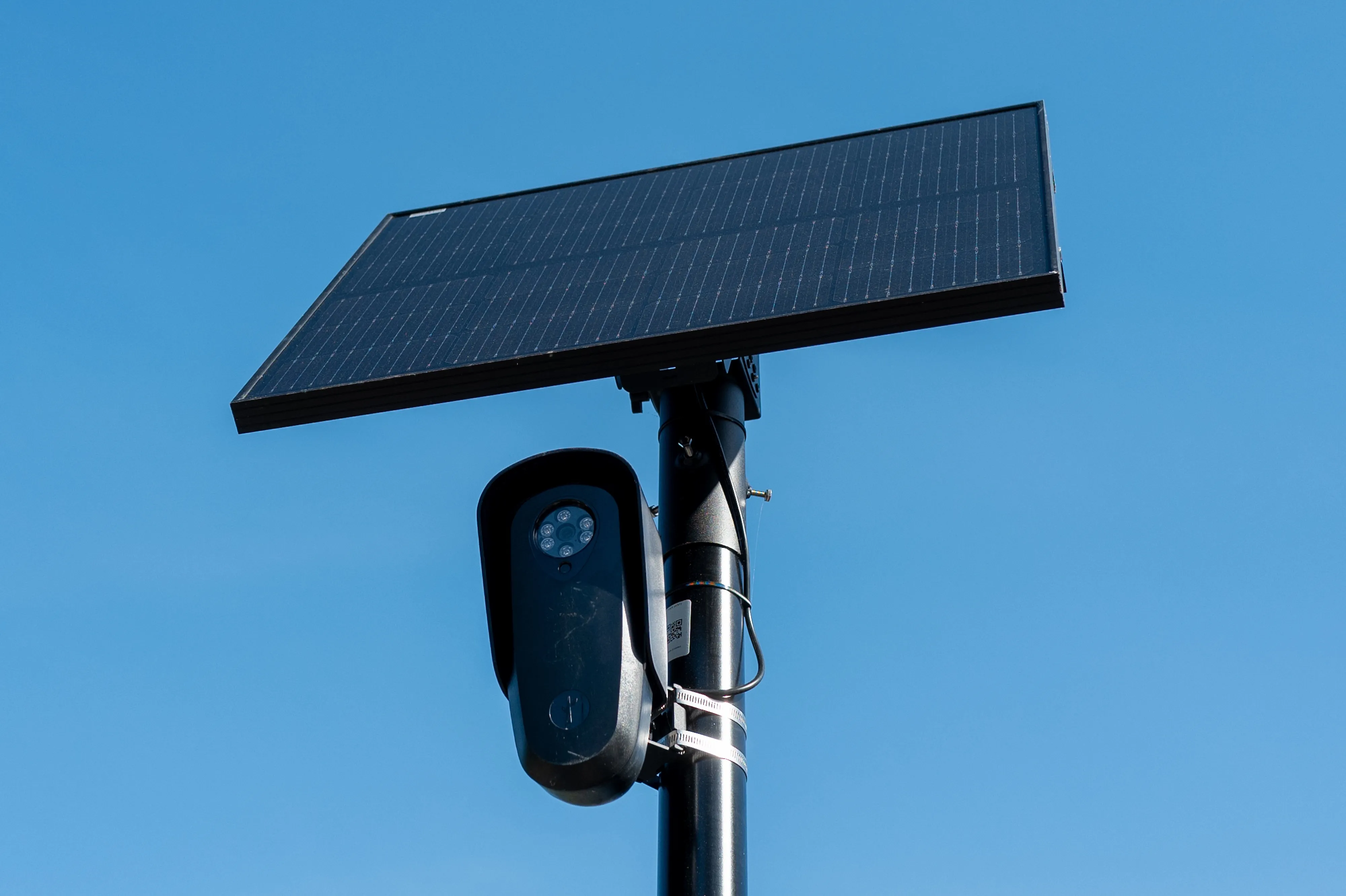A security camera mounted beneath a solar panel on a pole against a clear blue sky.