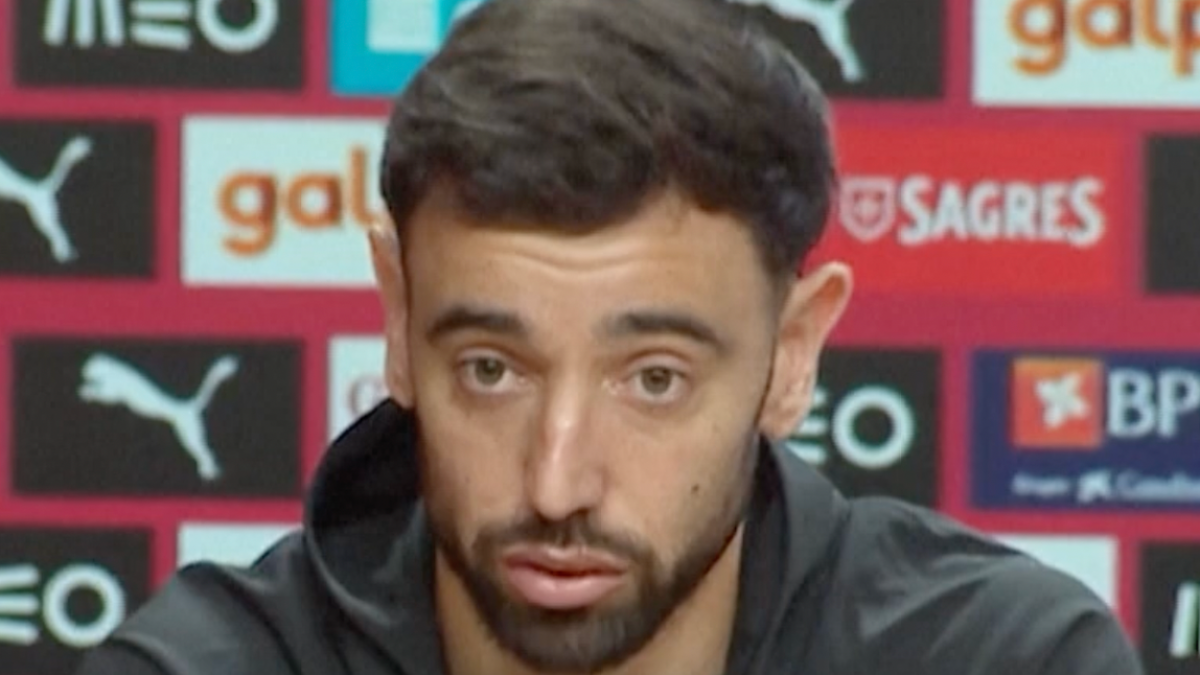 A bearded man in a dark jacket speaks at a press conference before a sponsor backdrop.