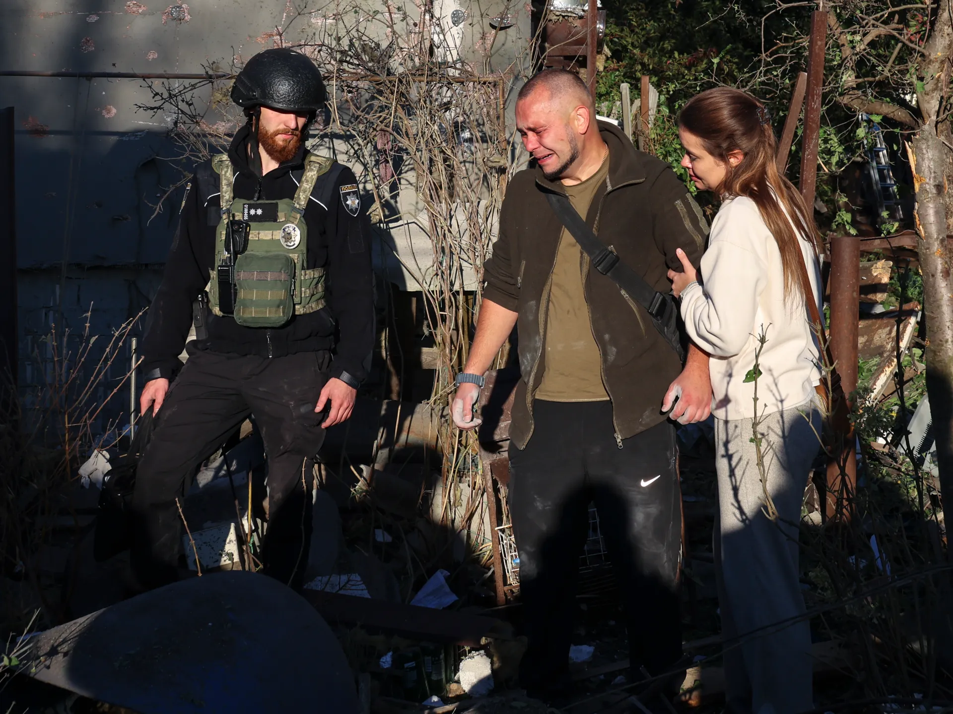 A distressed man walks beside a woman and an armed officer among debris and damaged structures.