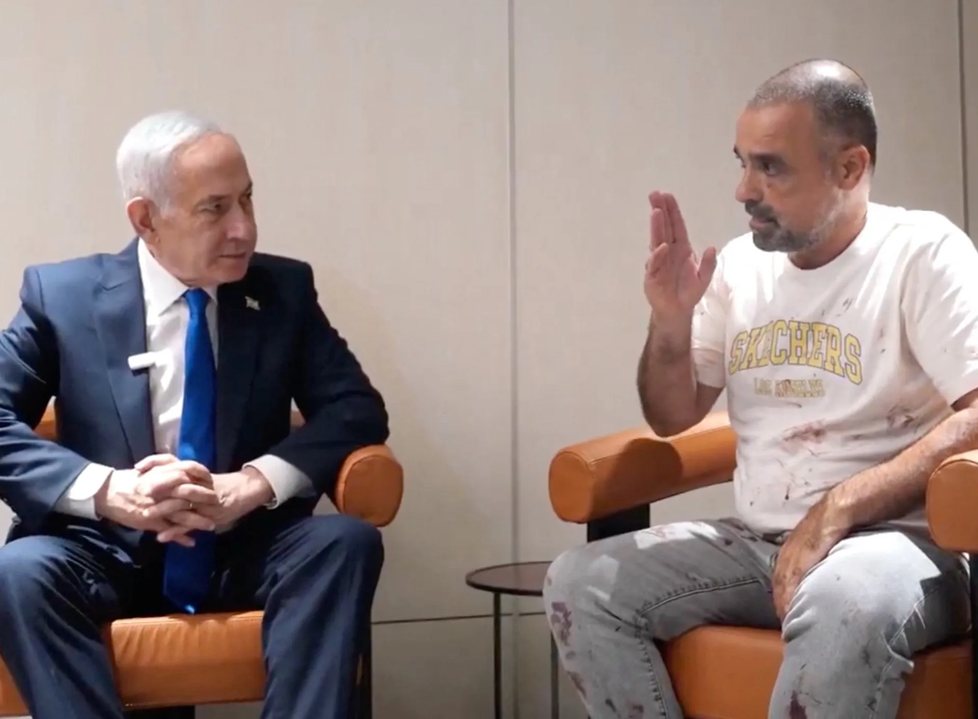 Two men sit facing each other; one wears a suit, while the other raises a hand while speaking.