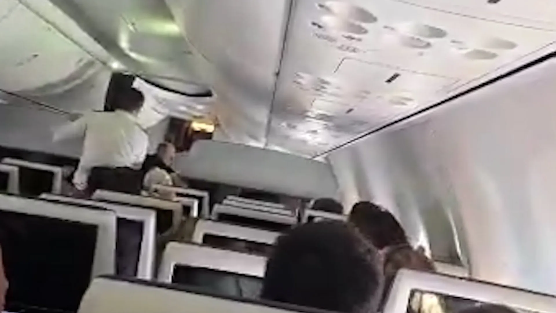 Passengers sit inside an airplane cabin as a person stands in the aisle near overhead bins.