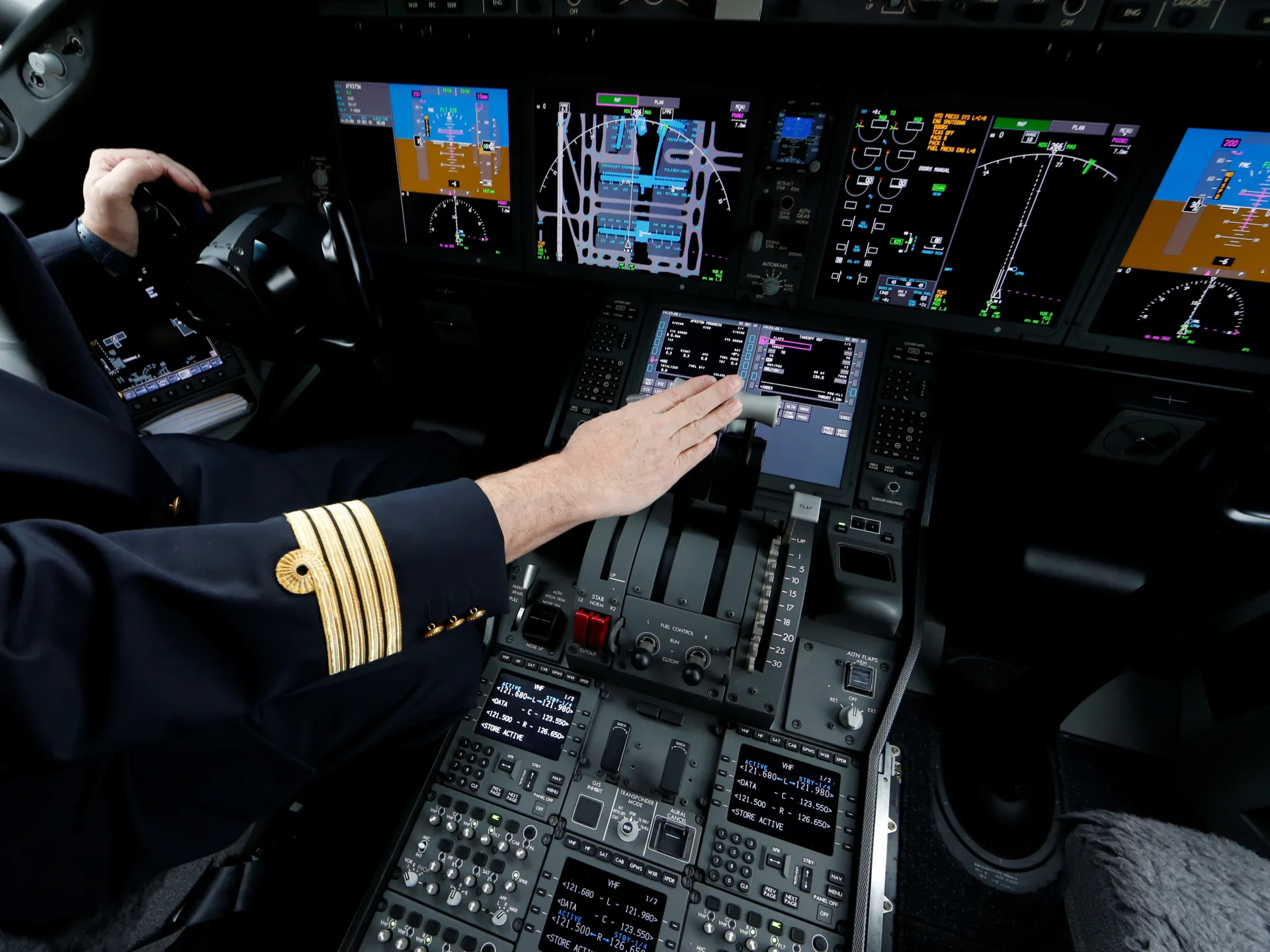 A pilot reaches across an aircraft cockpit control panel filled with illuminated flight displays.