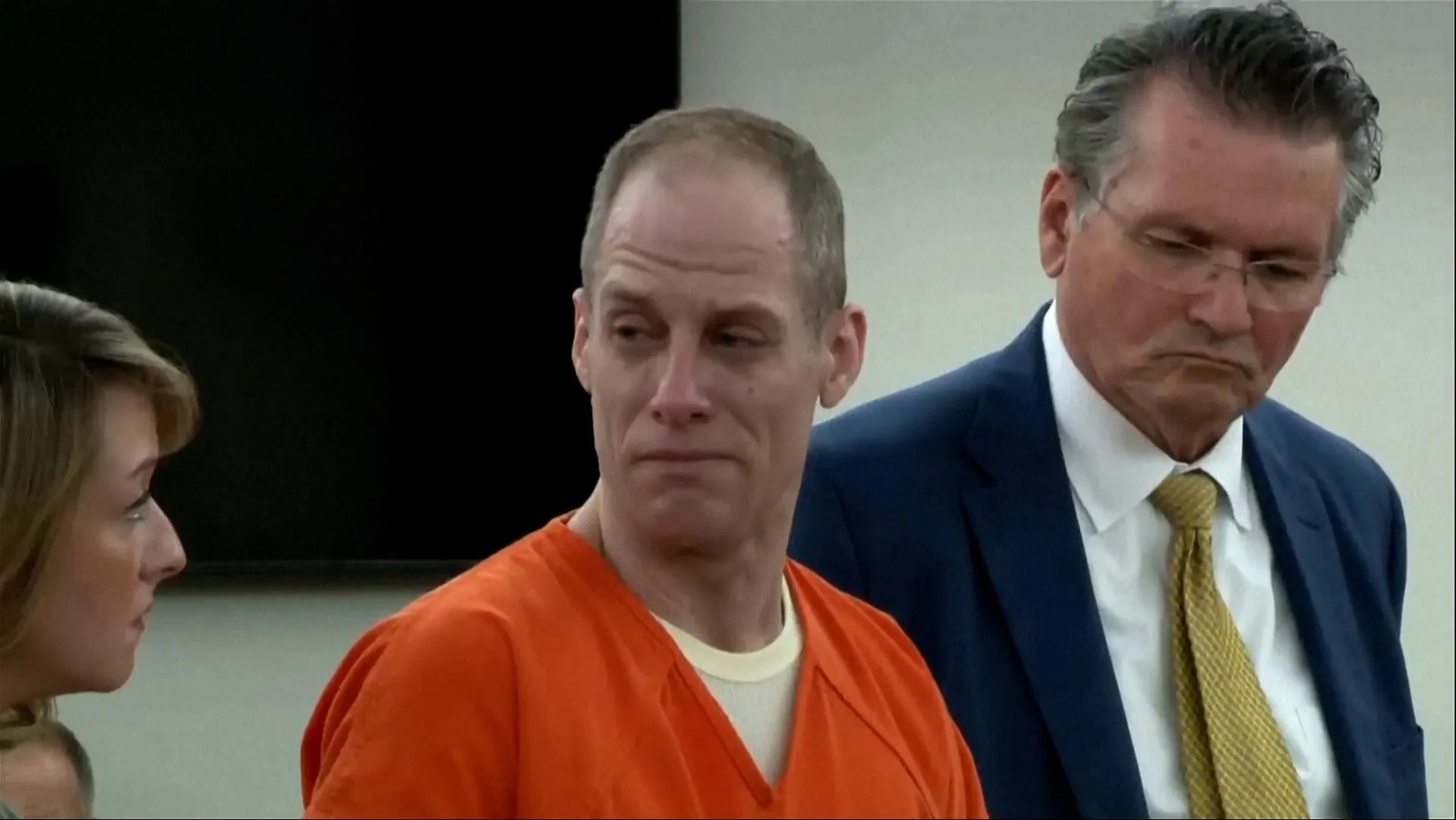 A man in an orange jail uniform stands between a woman and a suited man.