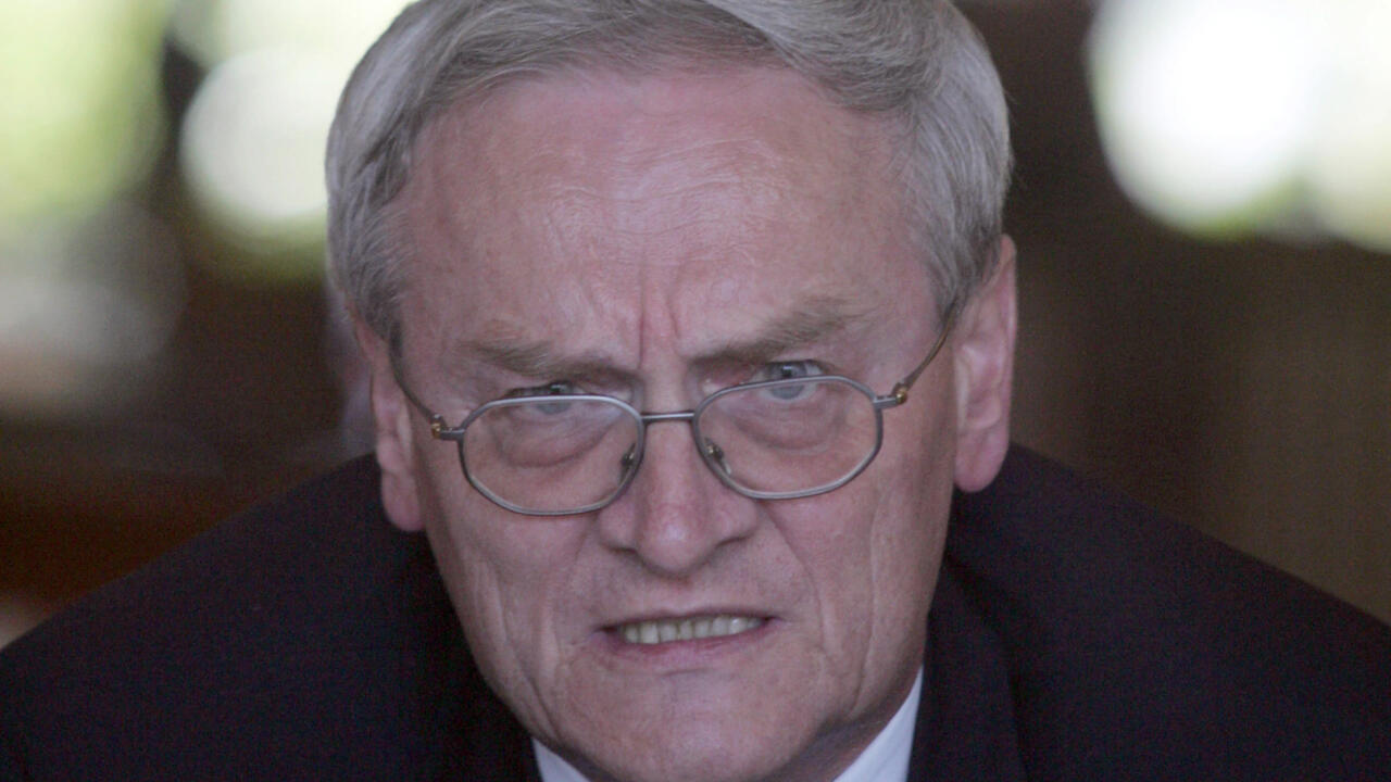 Close-up of an older man with gray hair and glasses, wearing a dark suit and looking forward.