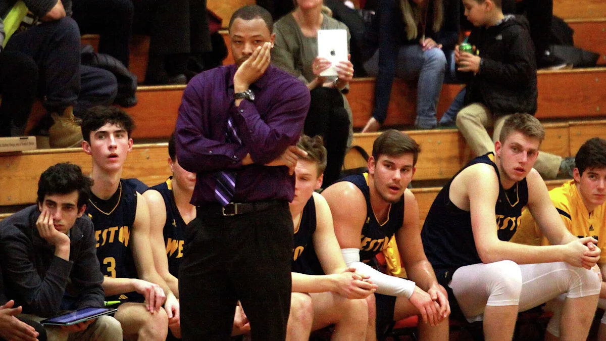A coach stands with arms crossed in front of seated basketball players during a game.