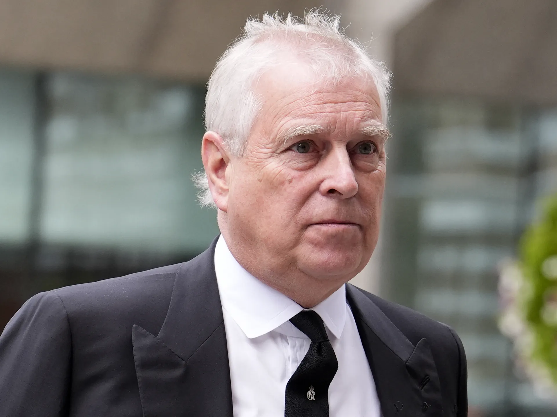 Older white-haired man in a dark suit and tie, pictured in close-up against a blurred background.