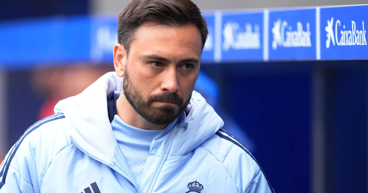 Bearded man in a pale blue football jacket stands before blue sponsor boards.