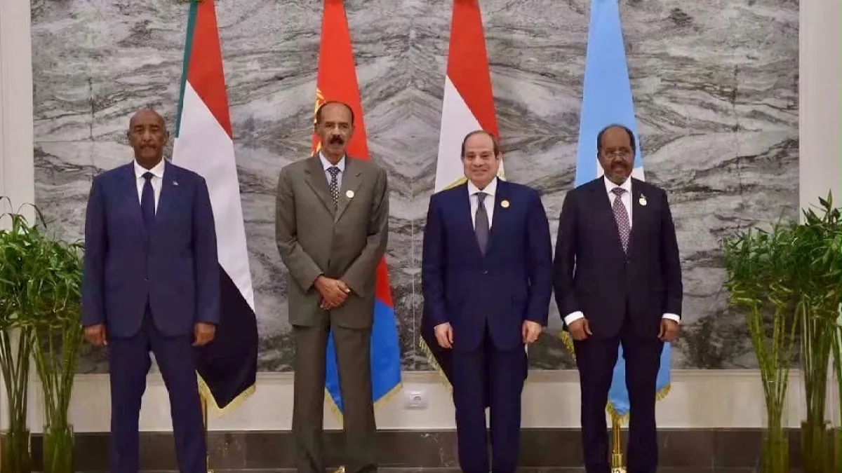 Four suited men stand in a row before national flags and a marble wall.