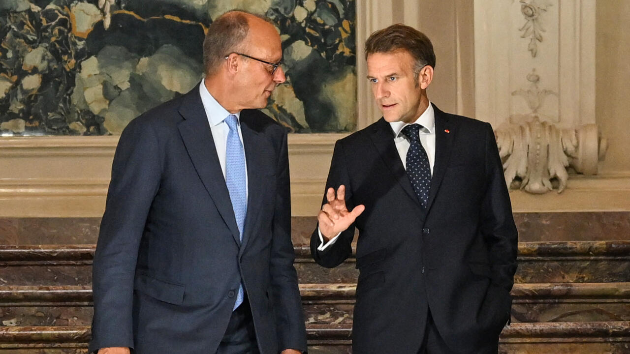 Two suited men talk indoors, one gesturing with his raised hand beside a marble staircase.
