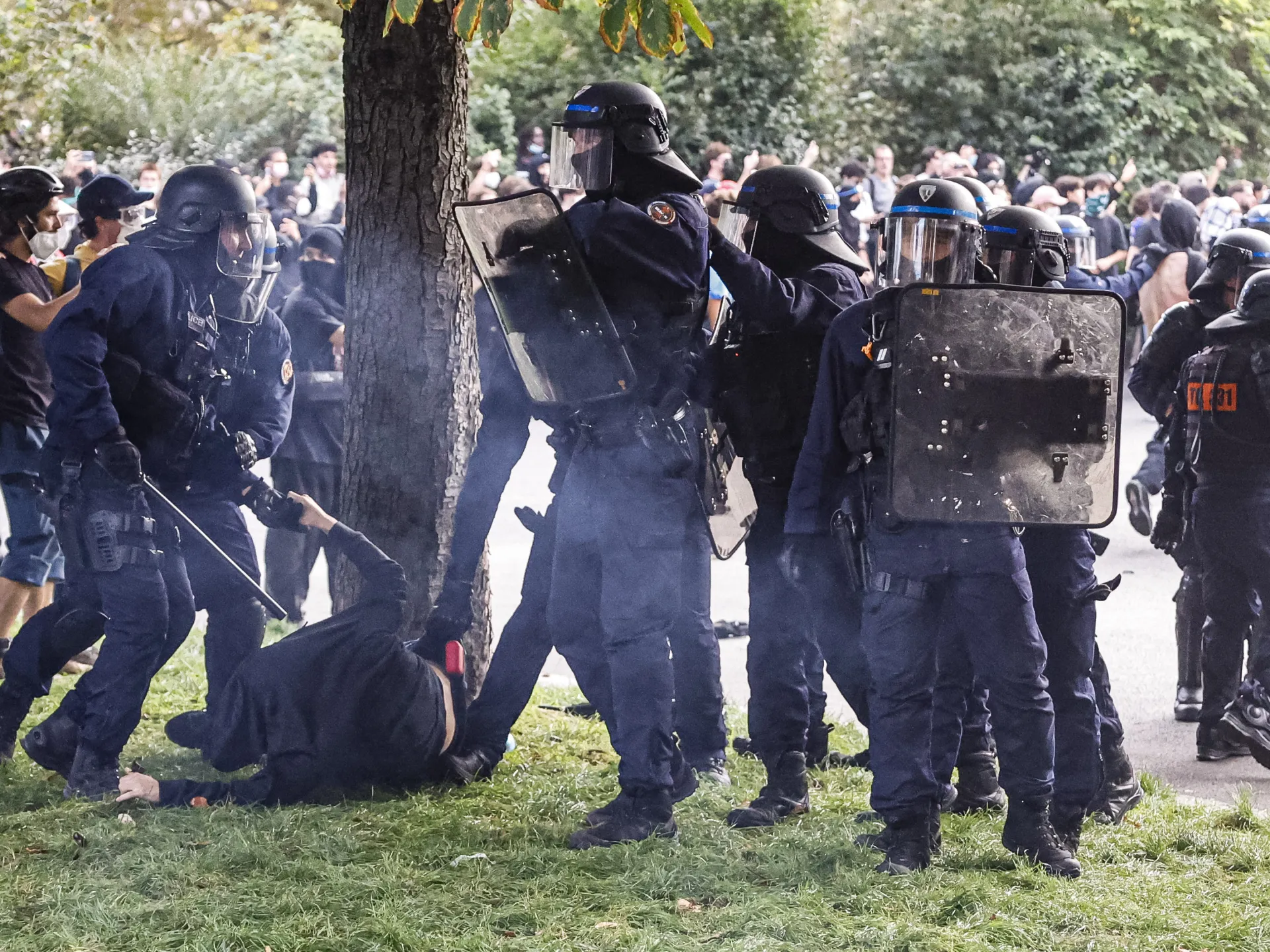 Helmeted police officers with shields stand around a person on the grass as a crowd gathers nearby.