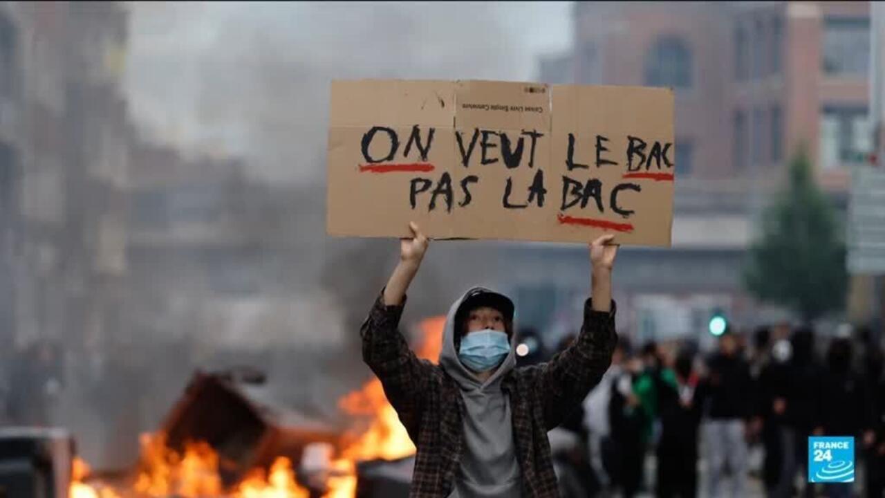 Masked person holds a handwritten cardboard sign above a smoky street as flames burn nearby.