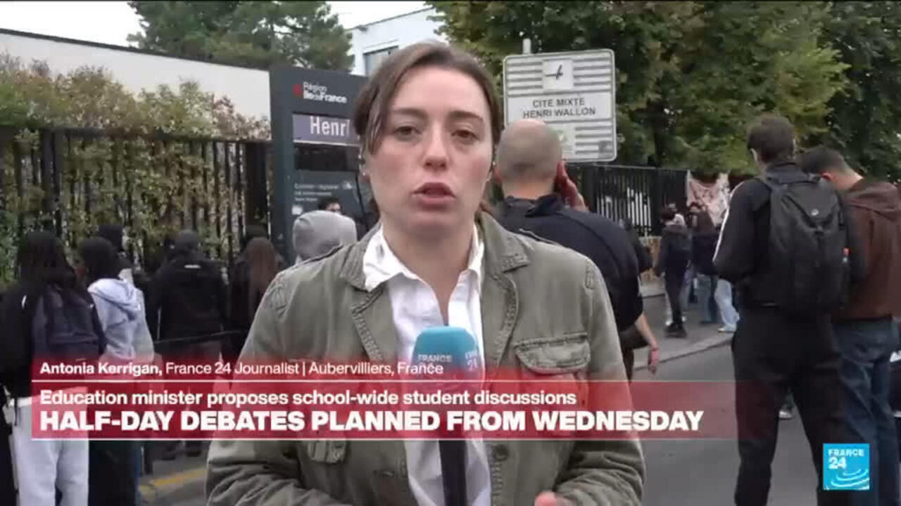 A reporter speaks outside a school as students and adults gather behind her near a gated entrance.