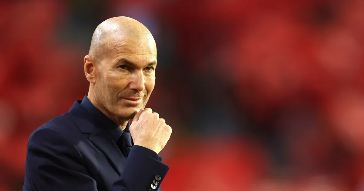 Bald man in a dark suit rests his hand against his chin against a blurred red background.
