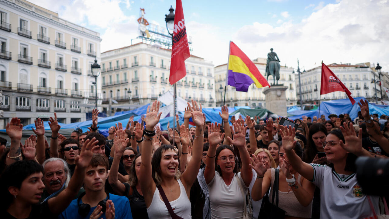 Crowd raises hands beneath protest flags in a city square lined with multi-story buildings.