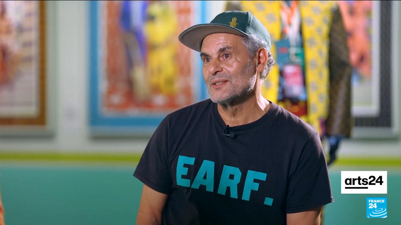 A man in a dark cap and black T-shirt sits before colourful framed artwork, with Arts24 and France 24 logos visible.