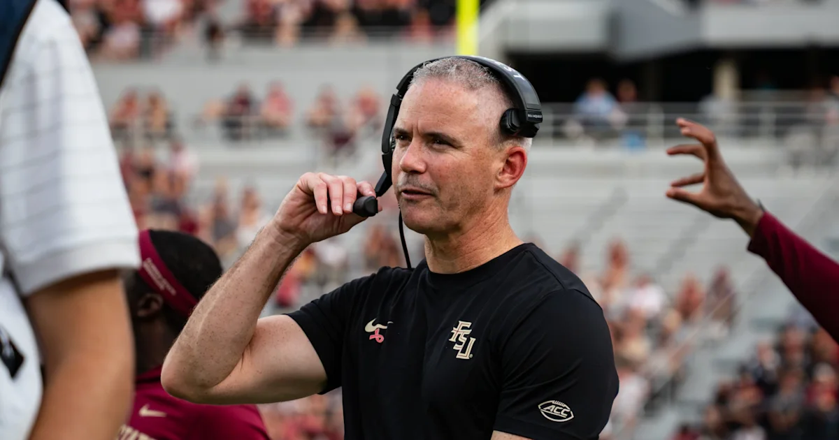 A football coach wearing a headset speaks into its microphone on the sideline during a game.