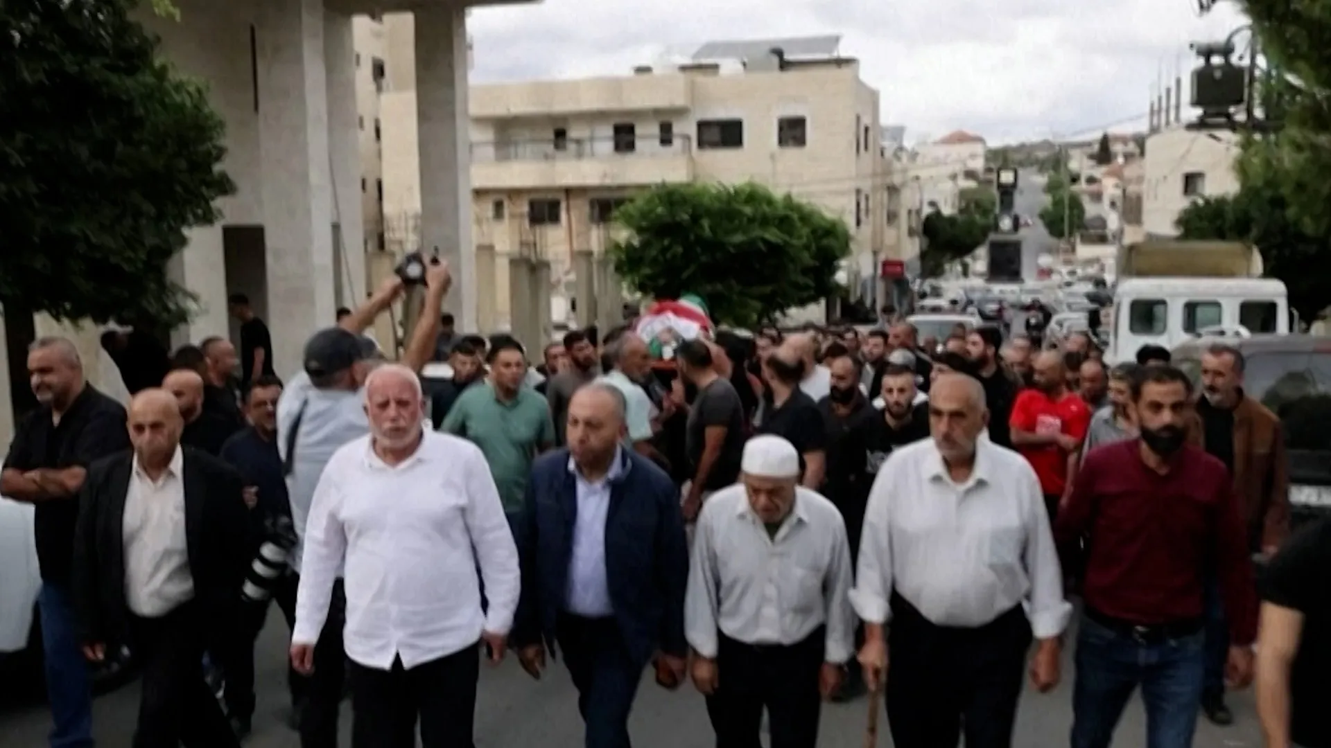 Men walk in a crowded street procession, with one person carrying a Palestinian flag.