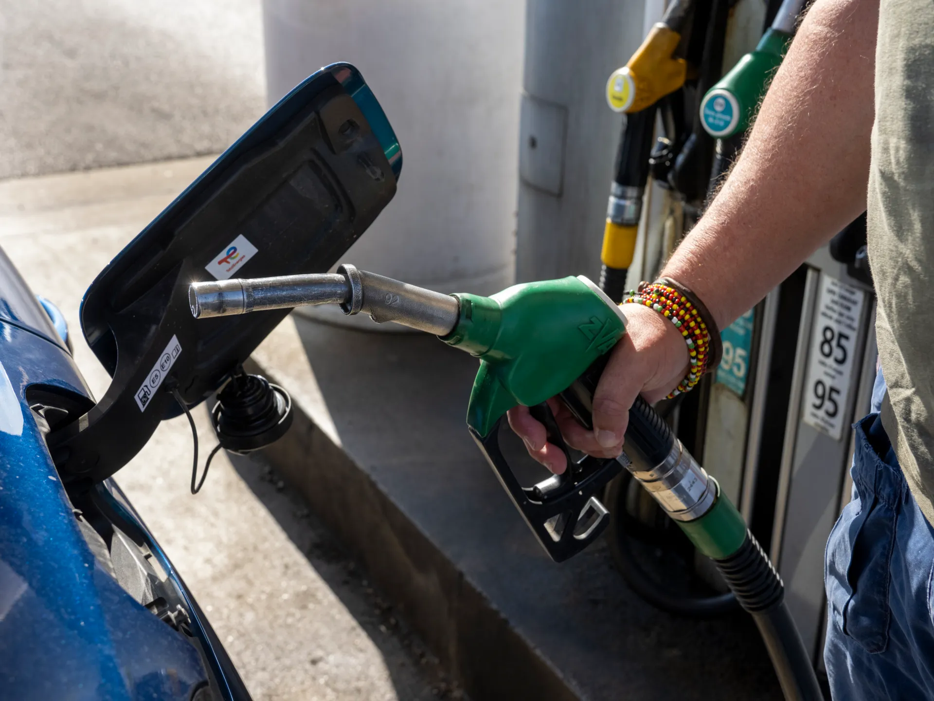 A person holds a green fuel nozzle beside a blue car at a gas station.